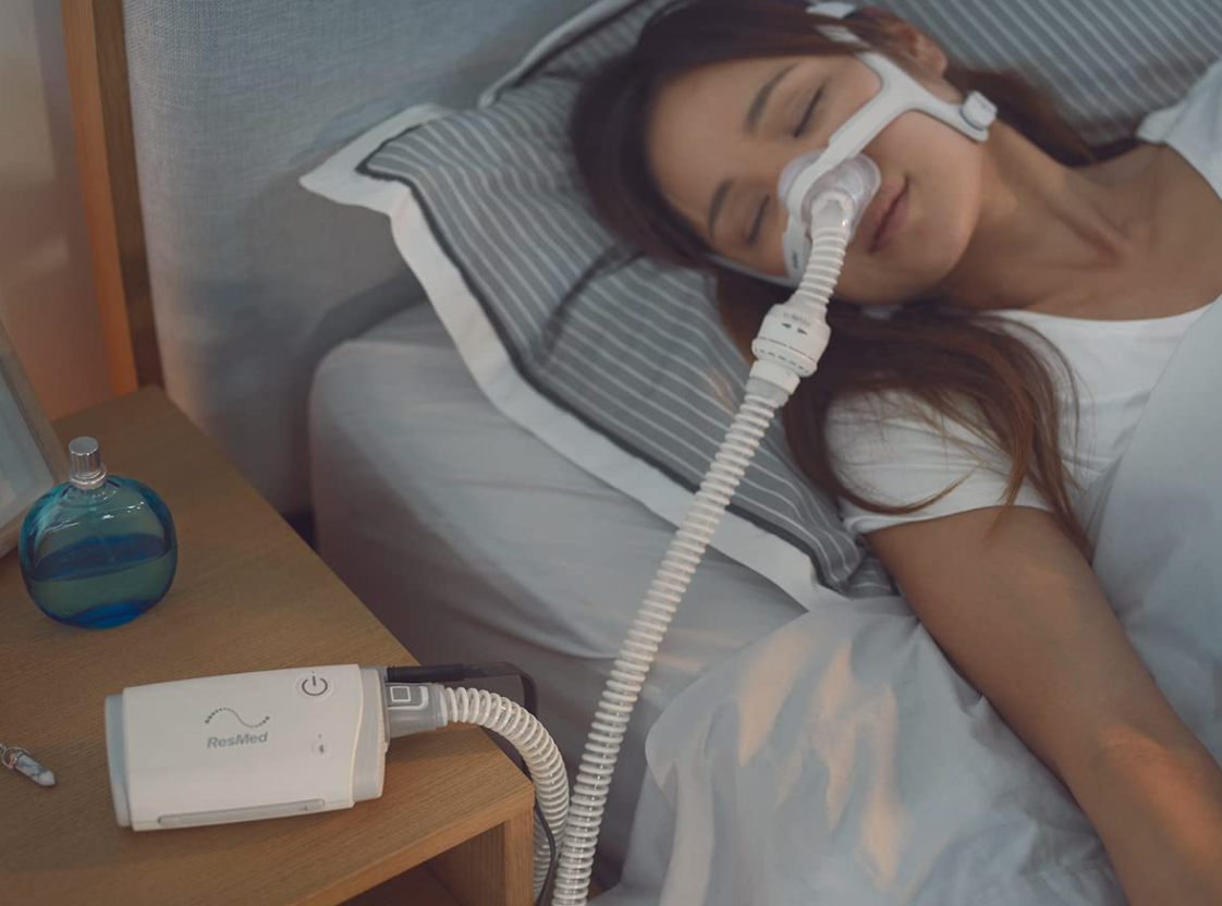 ResMed AirMini Portable Autoset CPAP device on bedside table with woman sleeping using nasal mask